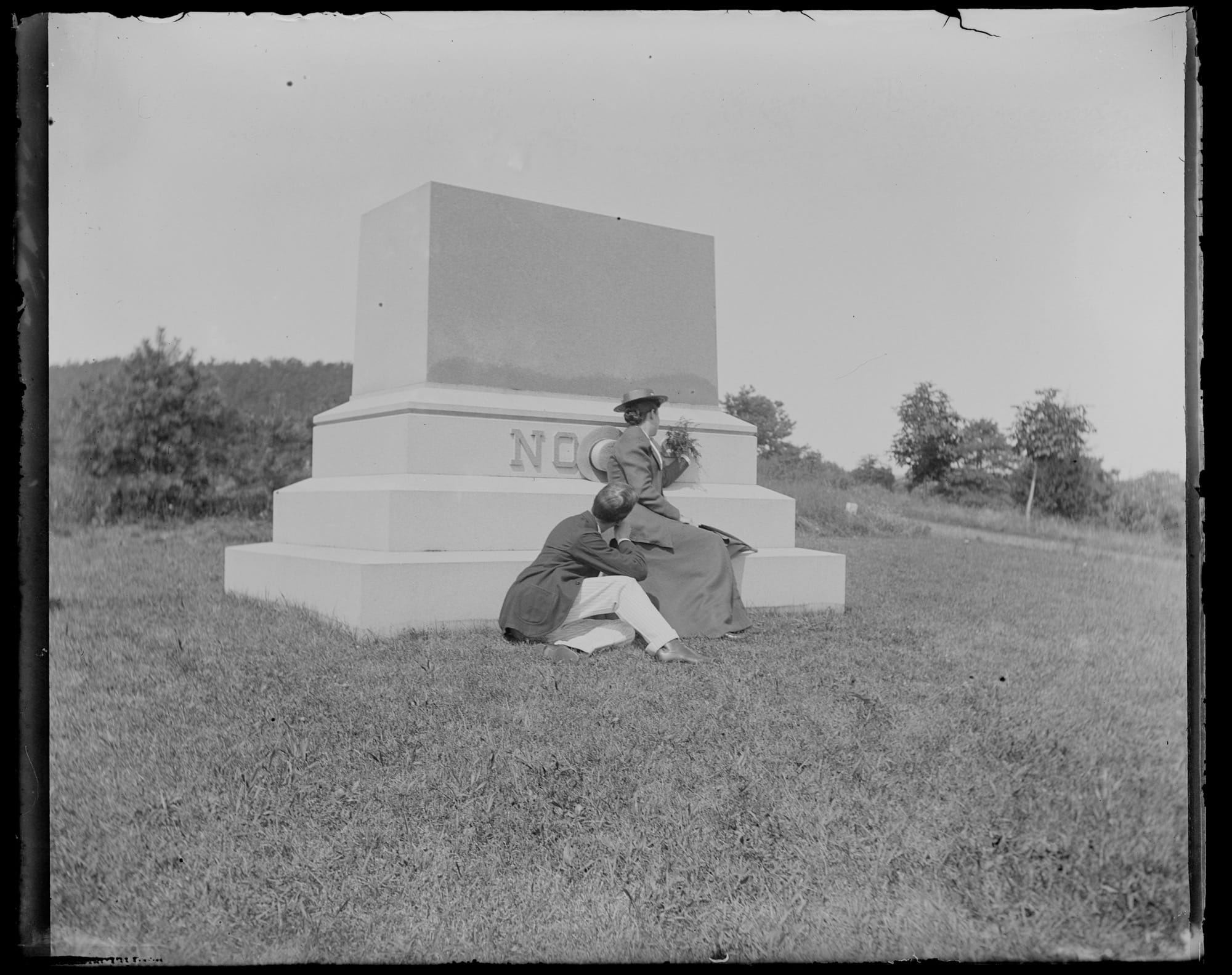 a black-and-white photo from the late 19th century of a man and woman seated at the base of a memorial, appearing to be getting engaged, with the word "NO" on the monument