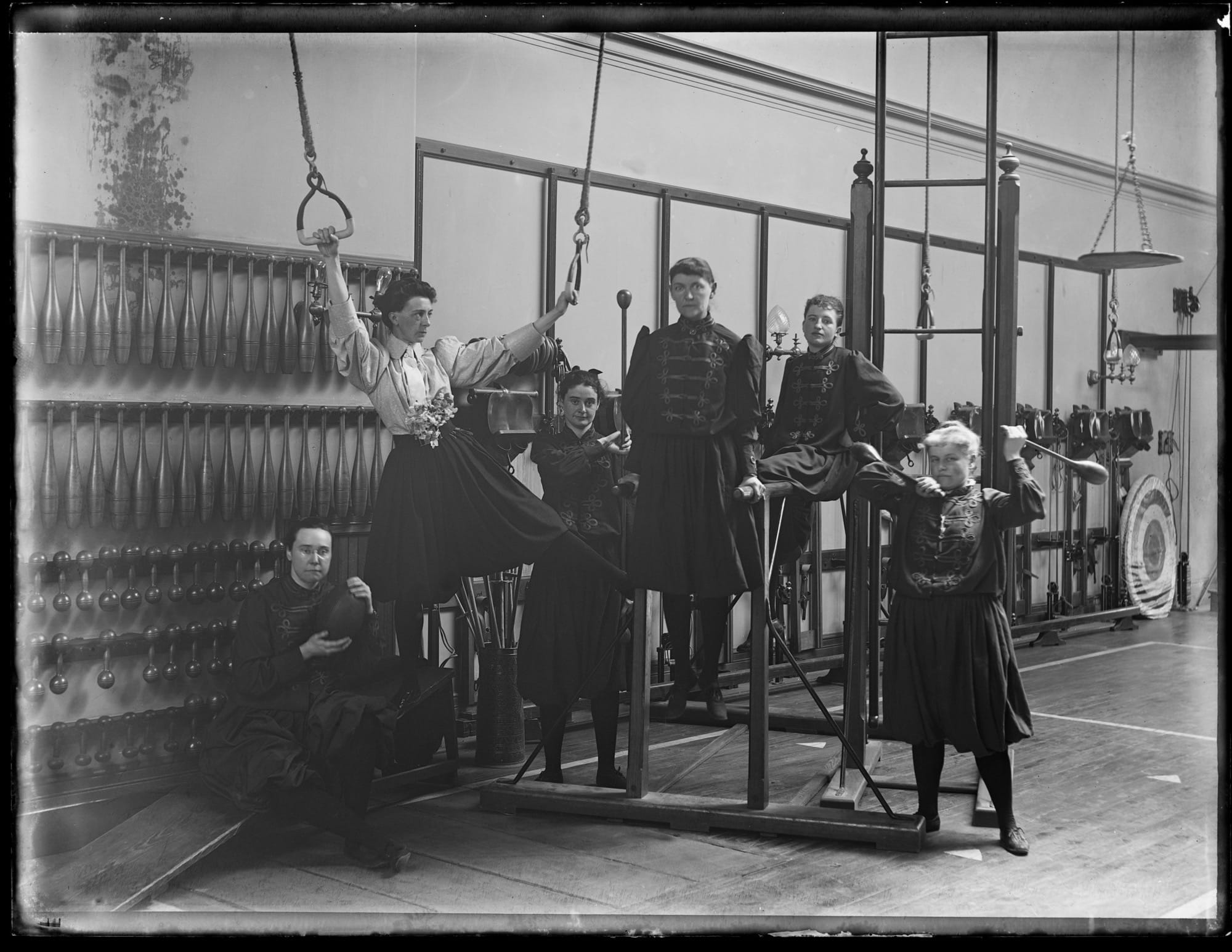 a black-and-white photo from the late 19th century of a group of young people posing around some exercise equipment in a gym
