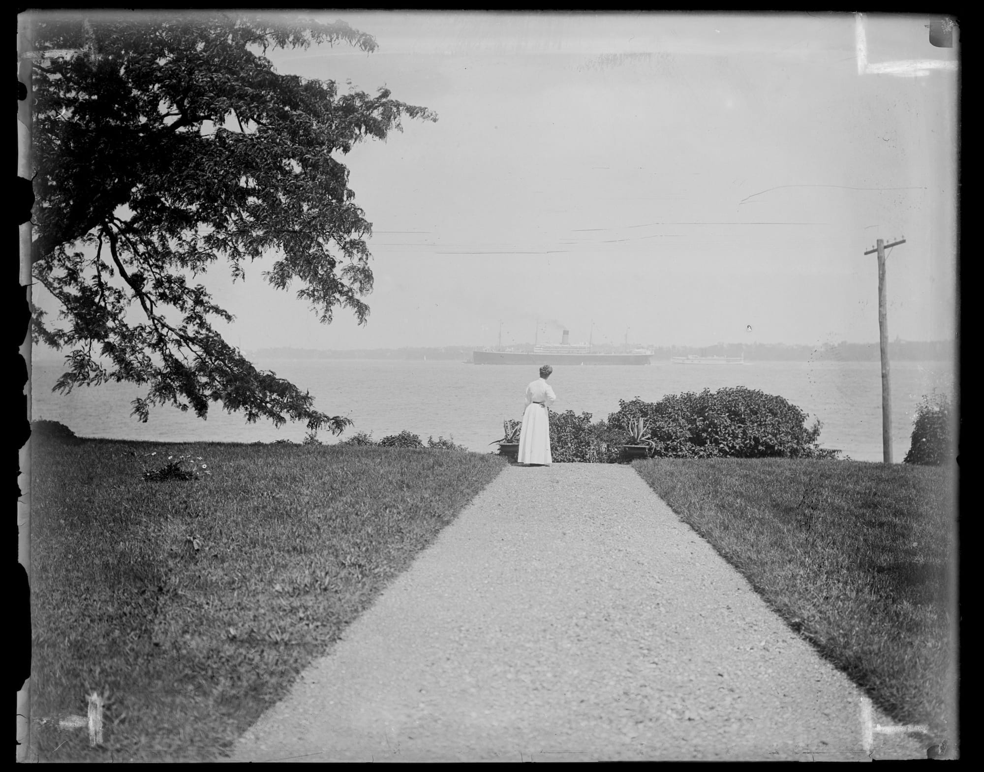 a black-and-white photo from the late 19th century of a woman in a white dress at the end of a path next to some water, where a large steam ship is out toward the horizon