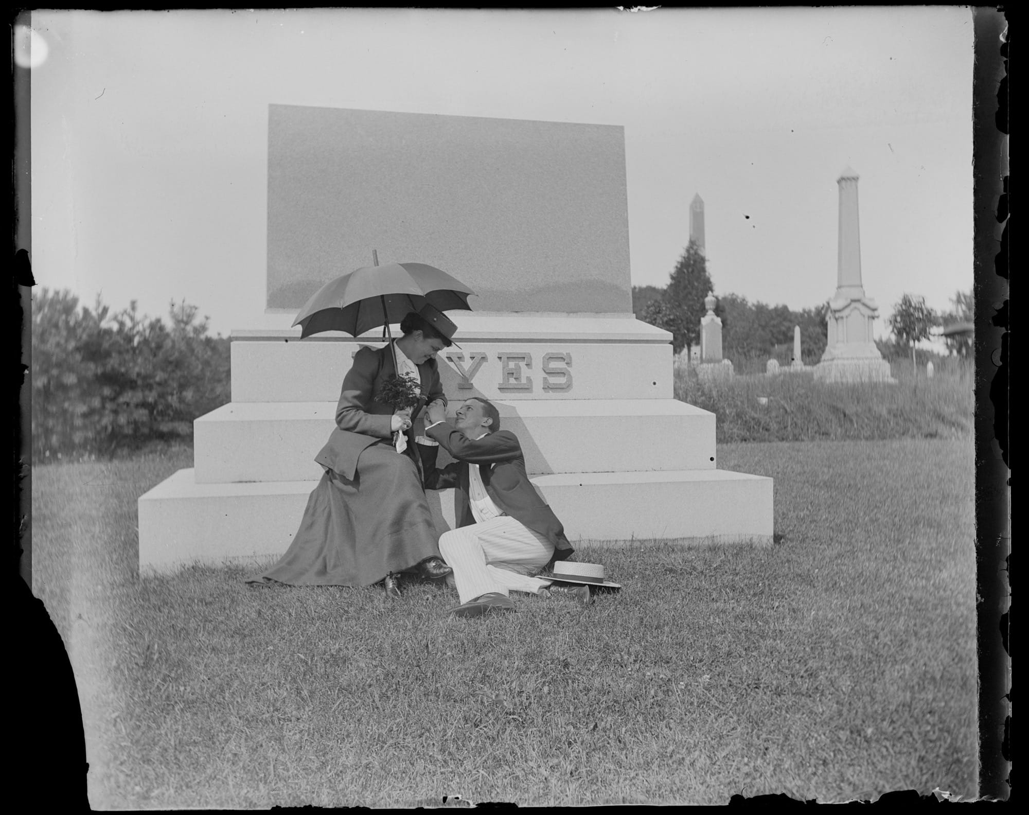 a black-and-white photo from the late 19th century of a man and woman seated at the base of a memorial, appearing to be getting engaged, with the word "YES" on the monument
