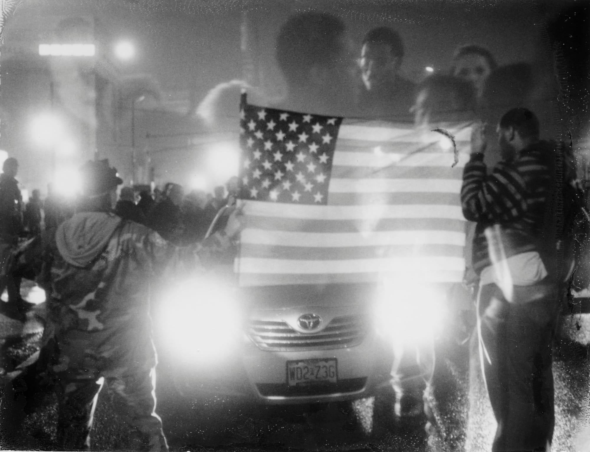 A black-and-white photograph by Carlos Javier Ortiz of demonstrators at night with an American flag