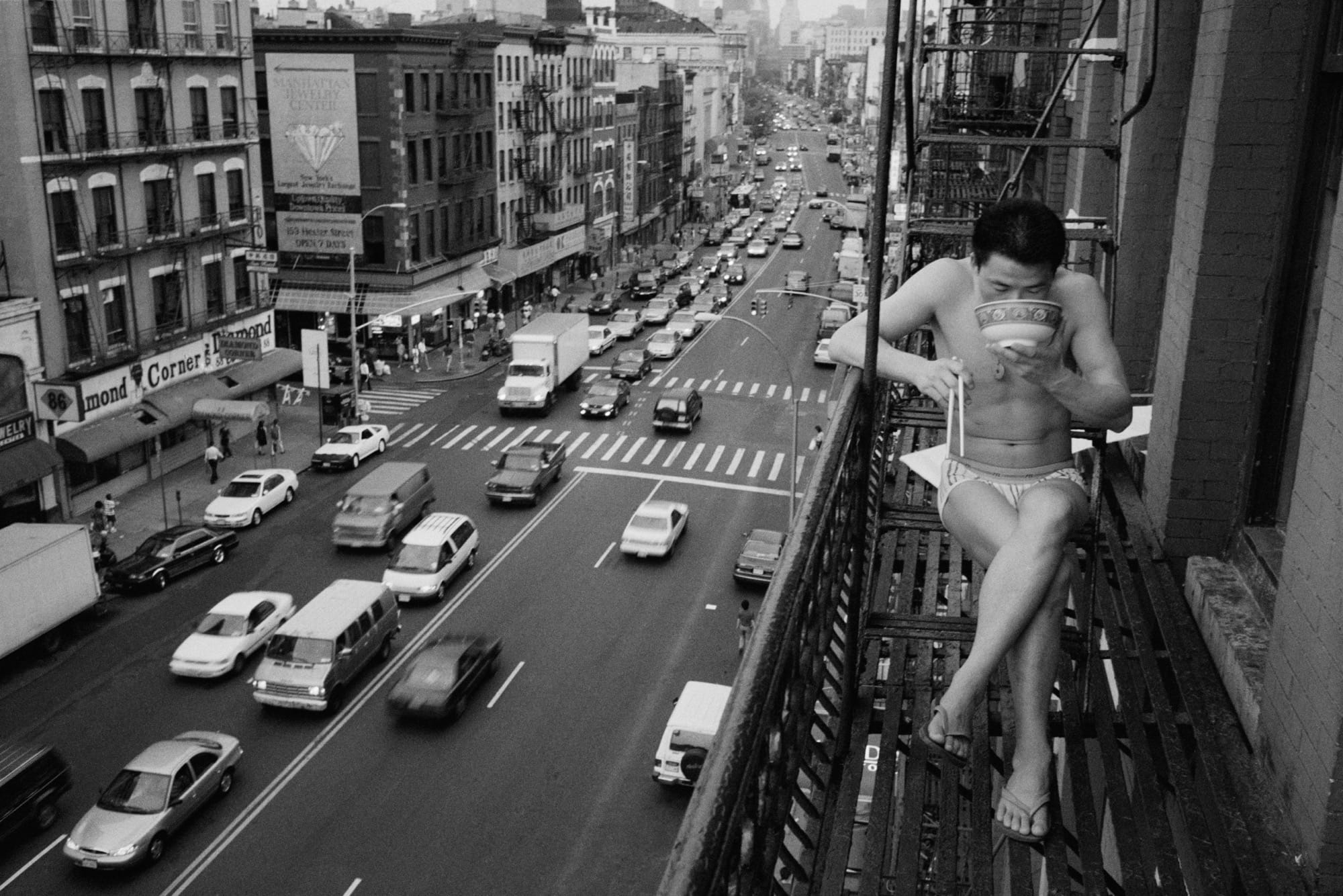 a black and white photo by Chien-Chi Chang of an asian man in his underwear sitting on a fire escape slurping noodles